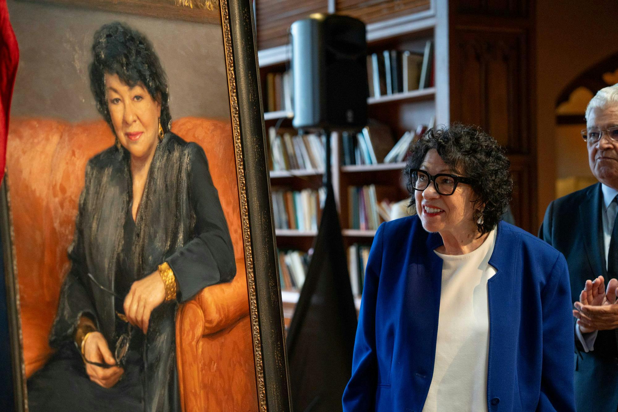 A woman with black curly hair, a royal blue jacket, and glasses smiles at a portrait depicting her.